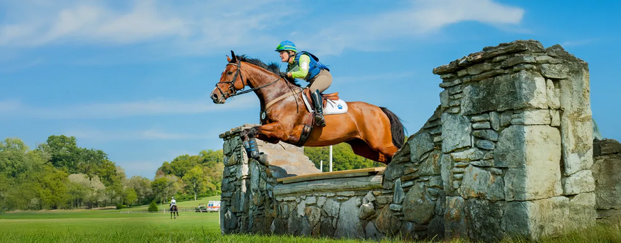 A rider in equestrian gear jumping with his horse over a stone obstacle in the countryside.