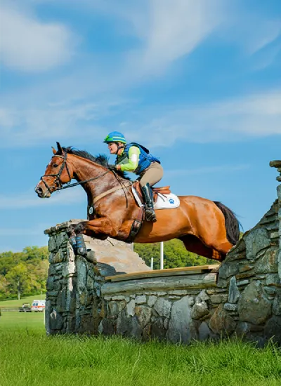 A rider in equestrian gear jumping with his horse over a stone obstacle in the countryside.