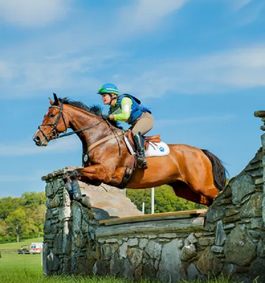 A rider in equestrian gear jumping with his horse over a stone obstacle in the countryside.
